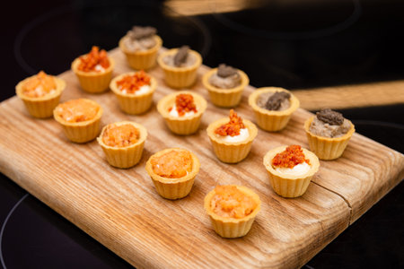 Mini savory tartlets with various colorful fillings like mousse and tapenade, arranged neatly on a wooden cutting board for catering.の写真素材