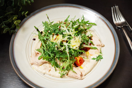 Close-up of a plated chicken salad topped with arugula, shredded cheese, cherry tomatoes, and balsamic glaze on a creamy base.の写真素材