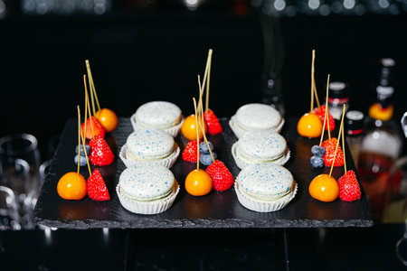 Elegant dessert display featuring blue-speckled macarons and colorful fruit skewers with strawberries, blueberries, and golden berries.の写真素材