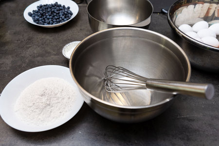Stainless steel mixing bowl with whisk surrounded by baking ingredients, including flour, eggs, and...