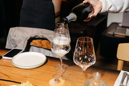 Close-up of a hand pouring sparkling wine into elegant glasses on a wooden table, set for a cozy and stylish dinner gathering.の写真素材