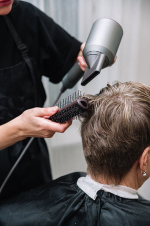 Professional hairstylist uses a round brush and blow dryer to style short hair in a salon during a grooming session.の写真素材