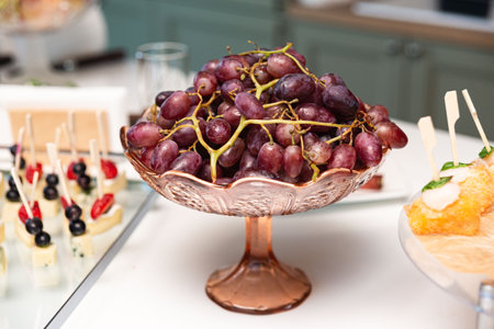 Bunch of ripe red grapes displayed in a decorative glass bowl on a white table, ideal for fruit platters or buffets.の写真素材