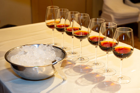 Several wine glasses are being filled with red wine on a white table, next to a metal bowl filled with ice cubes.の写真素材