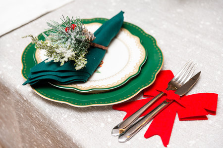 Festive holiday table setup with decorative green napkin, snow-dusted pine sprig, and red bow...