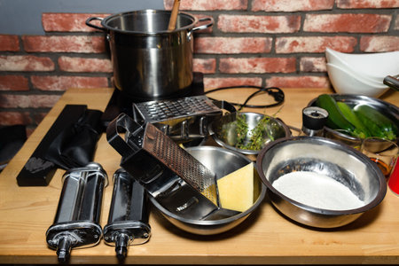 Kitchen scene showing cheese grater, pasta roller, flour, herbs, and vegetables on a wooden surface during meal prep.の写真素材