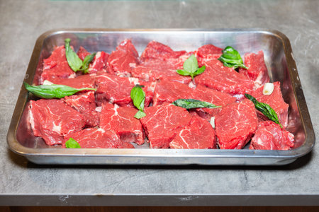 Fresh raw beef steak pieces arranged in a metal tray, garnished with basil leaves and garlic, prepared for marinating or cooking.の写真素材