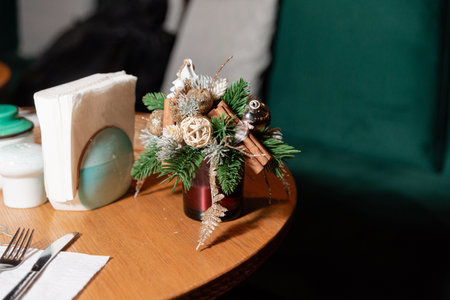 Small decorative Christmas arrangement featuring pine branches, cinnamon sticks, baubles, and glitter accents on a wooden dining table.の写真素材
