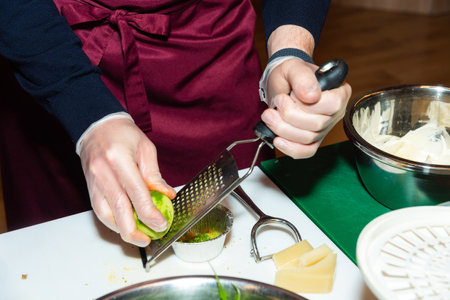 Close-up of gloved hands zesting a lime over a metal grater, with cheese, greens, and cooking tools...