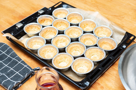 Freshly baked tart crusts in individual foil cups arranged on a parchment-lined baking tray, ready...