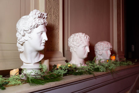 Three white marble-style busts displayed on a mantelpiece, surrounded by soft candlelight and green decorative foliage.の写真素材