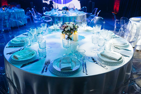 Luxurious round banquet table setup with crystal glassware, white linens, and floral centerpiece under blue ambient lighting.の写真素材