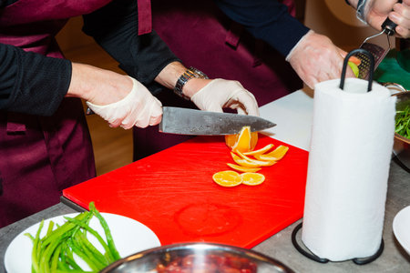 Close-up of chef's hands cutting a juicy orange on a red cutting board, with gloves and kitchen tools visible nearby.の写真素材