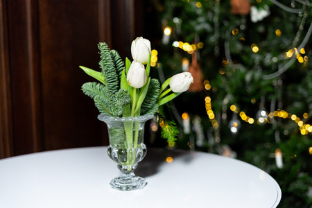 Elegant floral arrangement with white tulips and evergreen branches in a clear glass vase, placed on a round table during holidays.の写真素材