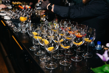 Bartender wearing gloves prepares cocktails behind a bar lined with empty stemmed glasses garnished with orange slices.の写真素材