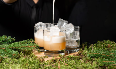 Elegant whiskey cocktails with ice and cream being poured, served on a rustic moss and pine-covered bar in a nature-inspired setting.の写真素材