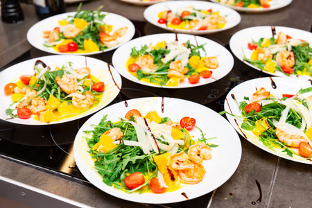 Multiple plates of fresh shrimp salad with arugula, cherry tomatoes, orange segments, cheese shavings, and balsamic drizzle on a counter.の写真素材