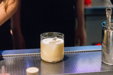 Close-up of a creamy yellow cocktail served in a lowball glass with ice cubes, placed on a metallic bar counter.の写真素材