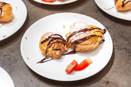 Two cream puffs topped with powdered sugar and chocolate sauce, served with fresh strawberry slices on a white plate.の写真素材