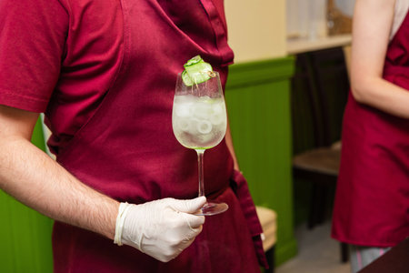 Person in red apron holding a clear cocktail with ice and cucumber garnish, likely at a drink-making workshop or bar event.の写真素材