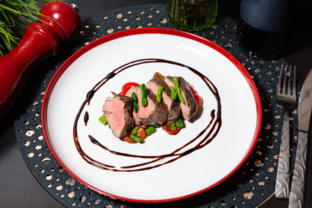 Sliced beef tenderloin served with asparagus and cherry tomatoes, artistically plated with balsamic reduction on a white dish.の写真素材