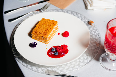 Elegant plated honey cake served with raspberry sauce, fresh berries, and edible flowers on a white plate at a formal event.の写真素材