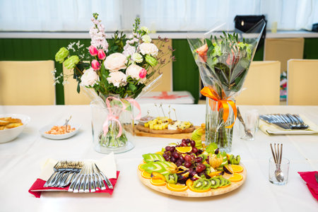 Beautifully arranged celebration table featuring fresh cut fruits, flower bouquets, utensils, and light snacks for a party or event.の写真素材