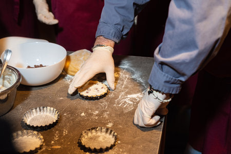 Gloved hands shaping mini tart crusts filled with cream and chocolate chips during a pastry-making process in a kitchen setting.の写真素材