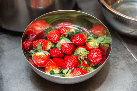 Bright red strawberries being cleaned in a metal bowl filled with water, preparing them for use in a fresh recipe.の写真素材