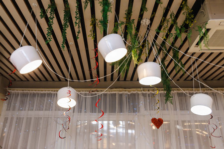 Decorative ceiling with white hanging pendant lights, green foliage, and red swirl ribbons creating a festive atmosphere indoors.の写真素材