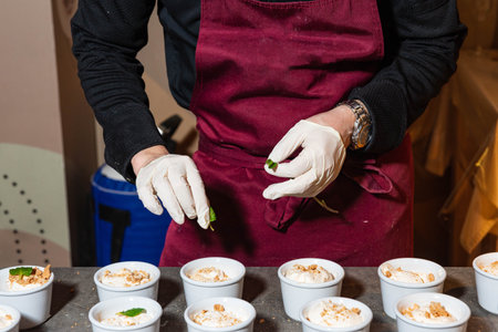 A chef in a burgundy apron adds mint leaves to creamy dessert cups with crumbled toppings, preparing...