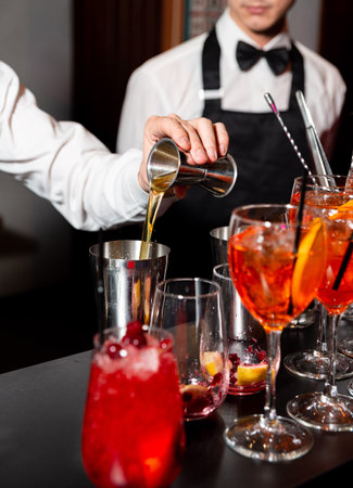 Bartender prepares cocktail by pouring liquid from jigger into shaker with vibrant drinks in the foreground at stylish bar.の写真素材