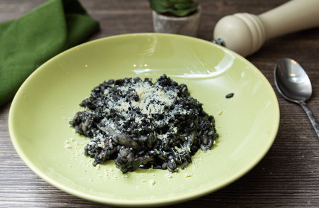 Rich black squid ink risotto topped with freshly grated Parmesan, served in a light green bowl on a wooden dining surface.の写真素材