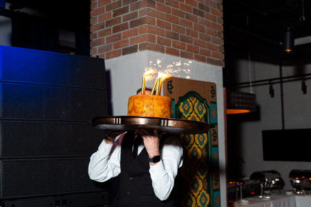 Waiter carries a festive cake topped with sparklers during a birthday or celebration event in a decorated venue.の写真素材