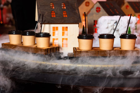 Paper coffee cups with lids and straws are placed on a model train, surrounded by fog, in a festive miniature village setup.の写真素材