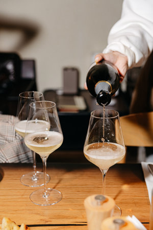 Close-up of a hand pouring sparkling white wine into tall wine glasses at a wooden dinner table during a social gathering.の写真素材