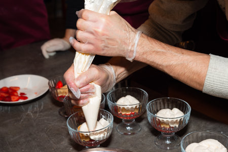 Close-up of hands using piping bag to fill dessert glasses with whipped cream over a strawberry base during food prep.の写真素材