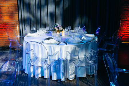 Elegant round table set for a formal event, featuring crystal glassware, white linens, candles, and transparent ghost chairs under moody lighting.の写真素材