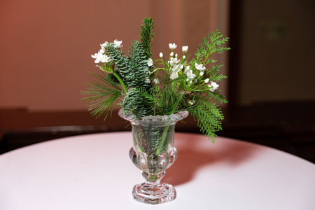 Elegant winter-themed bouquet with pine branches and white flowers in a clear glass vase on a round table.の写真素材