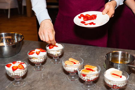 Close-up of a person garnishing layered dessert cups with fresh sliced strawberries and cream in a kitchen setting.の写真素材
