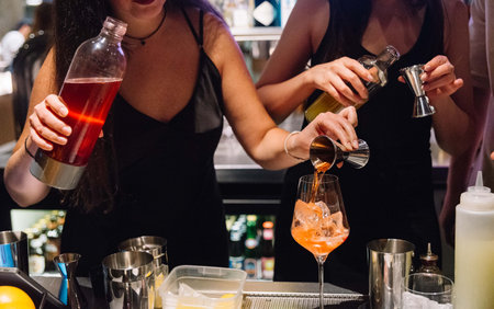 Two bartenders in black outfits preparing vibrant cocktails at a bar, using jiggers and bottles for precise mixing.の写真素材