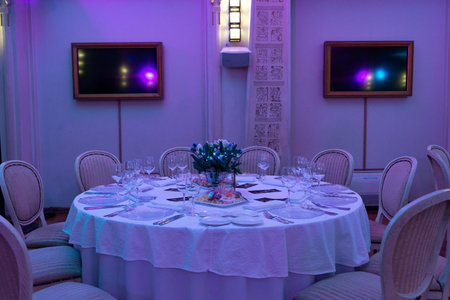 Round table set for a formal dinner with white tablecloth, wine glasses, cutlery, and a holiday-themed floral centerpiece.の写真素材