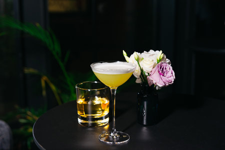 Two stylish cocktails placed beside a small floral arrangement on a black table in a dimly lit, modern setting.の写真素材