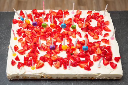 Large rectangular birthday cake topped with fresh strawberries and whipped cream, decorated with colorful candles for celebration.の写真素材