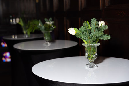 Glass vases with white tulips and pine sprigs decorate modern white-top cocktail tables in a dark, elegant venue setting.の写真素材
