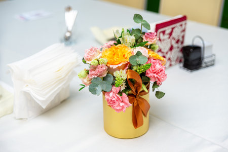 Bright flower arrangement in a gold pot with ribbon, placed on a table set with napkins and cutlery in soft light.の写真素材