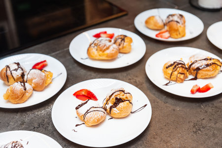 Dessert plates with cream-filled profiteroles, chocolate drizzle, powdered sugar, and fresh strawberry slices served on white plates.の写真素材