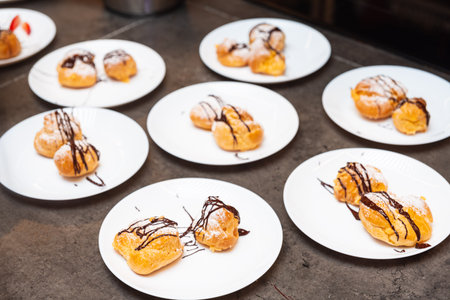 Plates of cream puffs topped with chocolate sauce and powdered sugar, arranged on a countertop for dessert service or event.の写真素材