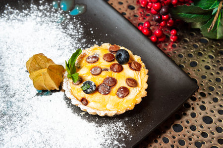 Close-up of a decorative dessert tart with blueberries, chocolate chips, and mint on a dusted plate, holiday-themed background.の写真素材