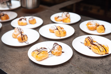 Multiple plates of cream puffs drizzled with chocolate sauce and dusted with powdered sugar, ready for serving or dessert buffet.の写真素材
